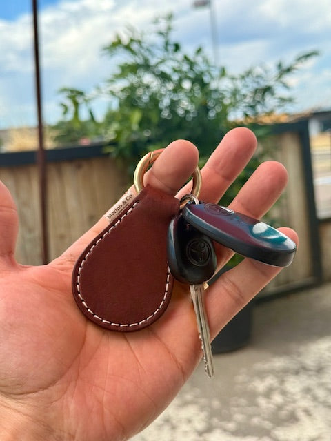 Hand holding a keychain with a leather tag and key fob against a blurred outdoor background