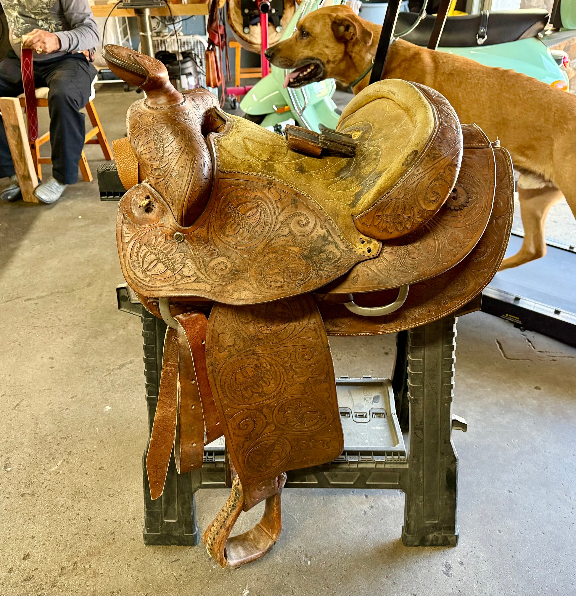 Brown leather saddle with intricate designs on a stand, with a dog in the background.